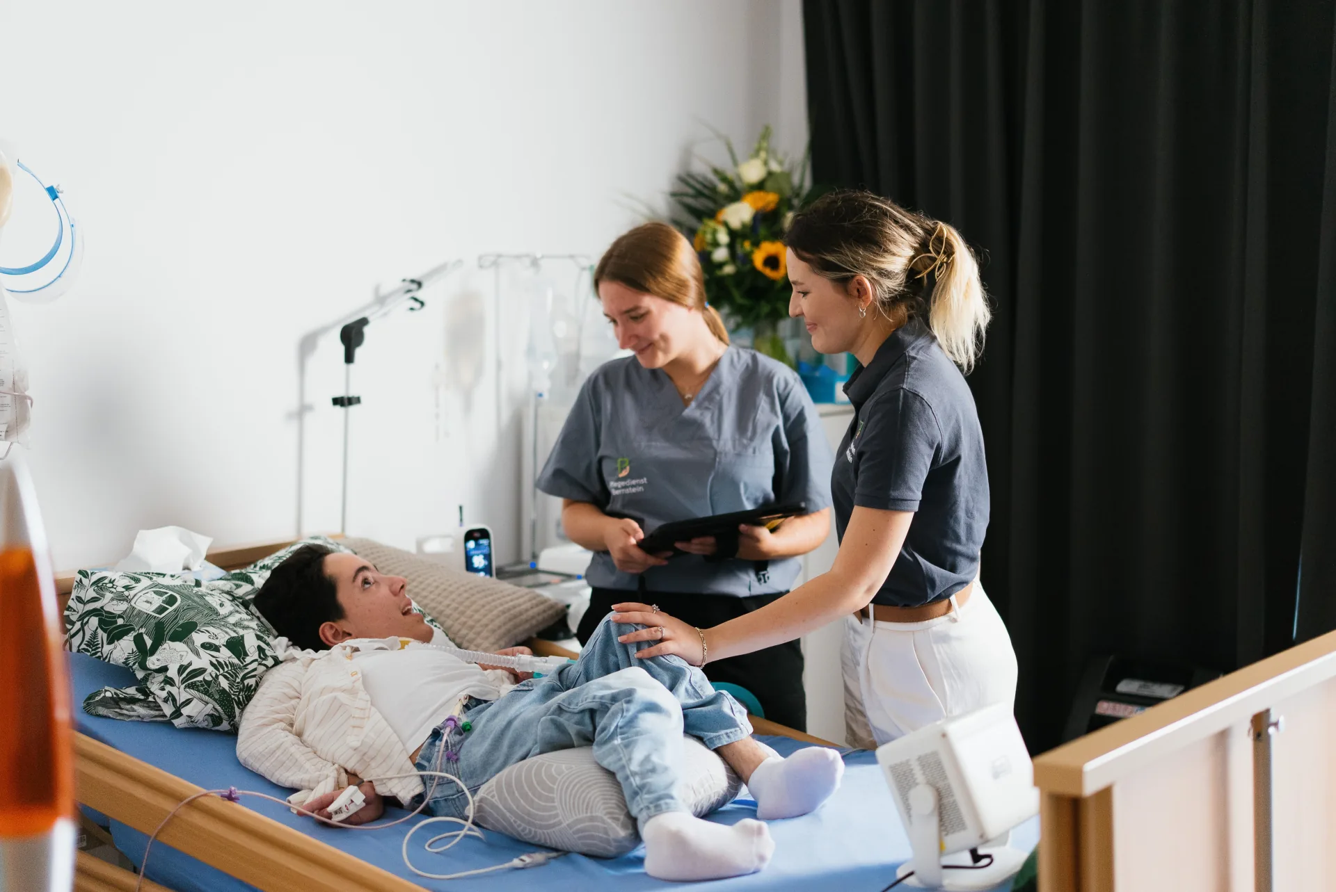 Zwei Pflegekräfte von Bernstein am Pflegebett eines beatmeten jungen Patienten, eine hält ein Tablet, die andere legt ihm die Hand aufs Knie.
